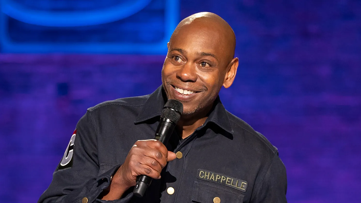 Dave Chappelle holding a microphone and smiling.